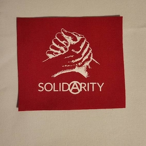 Puede incluir: Parche de tela rojo con una imagen blanca de manos entrelazadas y la palabra "SOLIDARITY" con un s&iacute;mbolo de anarqu&iacute;a en lugar de la "A".