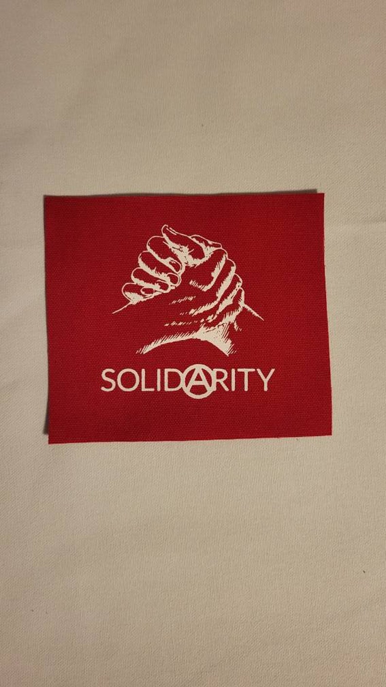 Puede incluir: Parche de tela rojo con una imagen blanca de manos entrelazadas y la palabra "SOLIDARITY" con un s&iacute;mbolo de anarqu&iacute;a en lugar de la "A".