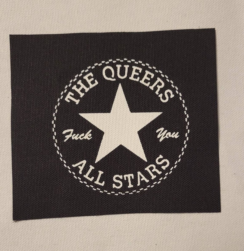 Op de afbeelding: Zwart vierkantige patch met een wit cirkelvormig ontwerp. Het ontwerp bevat een witte ster in het midden, omringd door de woorden "THE QUEERS" en "ALL STARS". De tekst "Fuck You" is ook in het ontwerp opgenomen.