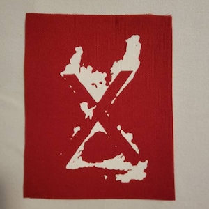 Op de afbeelding: Rode stoffen patch met een witte X in een verweerde stijl.