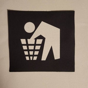 May include: A black and white patch with a stick figure throwing trash into a trash can. The patch is made of fabric and has a white outline.
