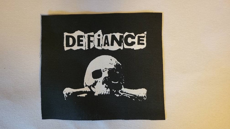 May include: Black and white embroidered patch with the word "DEFIANCE" in a distressed font and a skull and crossbones design.