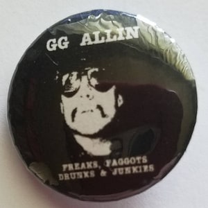 May include: Black and white button with a portrait of GG Allin wearing sunglasses. The text "GG ALLIN" is at the top of the button. The text "FREAKS, FAGGOTS DRUNKS & JUNKIES" is at the bottom of the button.