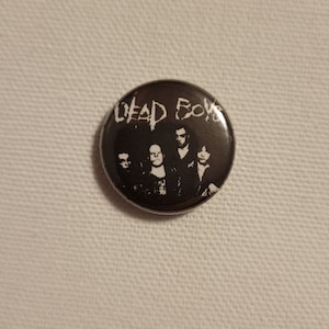 Pode incluir: Um botão preto e branco com o texto "Dead Boys" e uma imagem preto e branco de uma banda.
