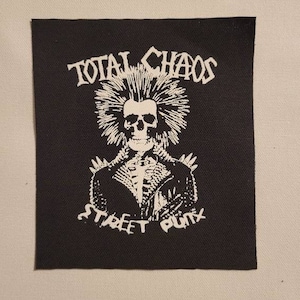 Peut inclure: Patch noir et blanc avec un crâne portant une veste en cuir et une crête iroquoise. Le texte "TOTAL CHAOS STREET PUNK" est autour du crâne.