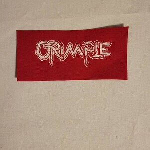 Op de afbeelding: Een rood stoffen patch met het woord "CRUMPLE" in witte, gestileerde letters.