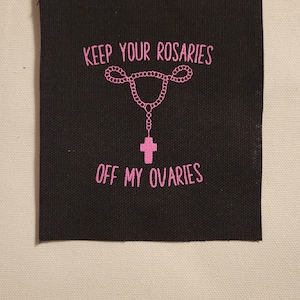 May include: Black fabric patch with pink text that reads "Keep your rosaries off my ovaries". A pink rosary with a cross is depicted above the text.