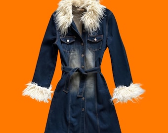 May include: A long-sleeved dark blue denim coat featuring a faded wash down the center, a button-down front, and a matching tie belt. The garment is accented with long, shaggy off-white faux fur trim along the large collar and the sleeve cuffs.