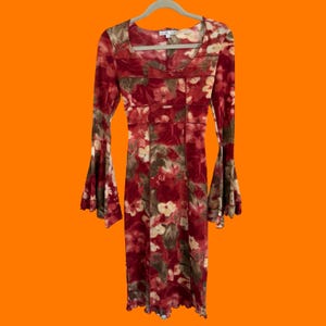 Puede incluir: Vestido largo hasta la rodilla con mangas largas y estampado floral en tonos rojos, marrones y crema. El vestido tiene un escote redondo y mangas acampanadas. Cuelga de una percha blanca sobre un fondo naranja.