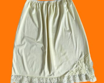 May include: A pale yellow half slip featuring a smooth, opaque fabric with an elasticized waistband. The hem is finished with a wide, scalloped white lace trim that extends upward in a triangular floral pattern on the lower right side.