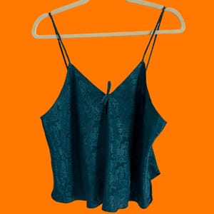 May include: Teal-colored camisole with a paisley pattern. The top features a V-neckline and thin spaghetti straps. A small bow detail is centered at the neckline. The camisole is hanging on a white hanger against an orange background.
