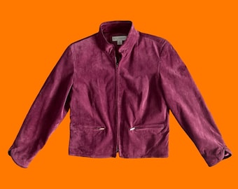 May include: A burgundy suede jacket with a zip-up front and two front pockets. The jacket has a stand-up collar and silver-toned zippers. The sleeves have a button detail at the cuffs. The jacket is displayed against an orange background.