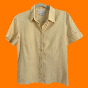 May include: A pale yellow short-sleeved shirt with a button-down front. The shirt features a collared neckline and is suspended on a hanger. The sleeves have a stitched detail. The shirt is made of a light-coloured fabric.