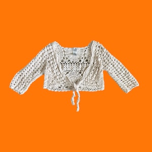 May include: A cream-colored, long-sleeved crochet shrug featuring an intricate openwork pattern, a scalloped edge, and a front tie closure. The garment includes a small white label at the inner neckline with the text LILY printed in black.
