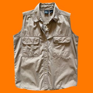 May include: A sleeveless, button-down shirt in a tan color. The shirt has two front pockets and a collared neckline. The brand label "BE" is visible on the collar. The shirt is made of a lightweight fabric.