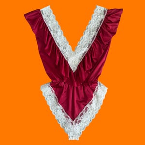 May include: Burgundy-colored bodysuit with white floral lace trim. The V-neckline and edges are adorned with delicate lace. The garment has a satin-like sheen and a ruffled design along the neckline.
