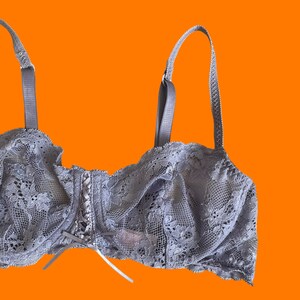 May include: A light gray lace bra with adjustable straps and a delicate floral pattern. The bra features a center bow and a scalloped edge. The background is a bright orange.