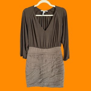 Puede incluir: Vestido marrón con mangas largas y cuello en V. La parte superior es de color marrón liso, mientras que la falda tiene un diseño plisado texturizado. El vestido está colgado en una percha blanca sobre un fondo naranja.