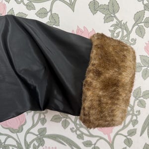 May include: Close-up of a black garment sleeve with a brown faux fur cuff. The sleeve is made of a shiny, smooth fabric. The faux fur cuff is a warm, light brown color, adding a touch of luxury. The background features a floral wallpaper.