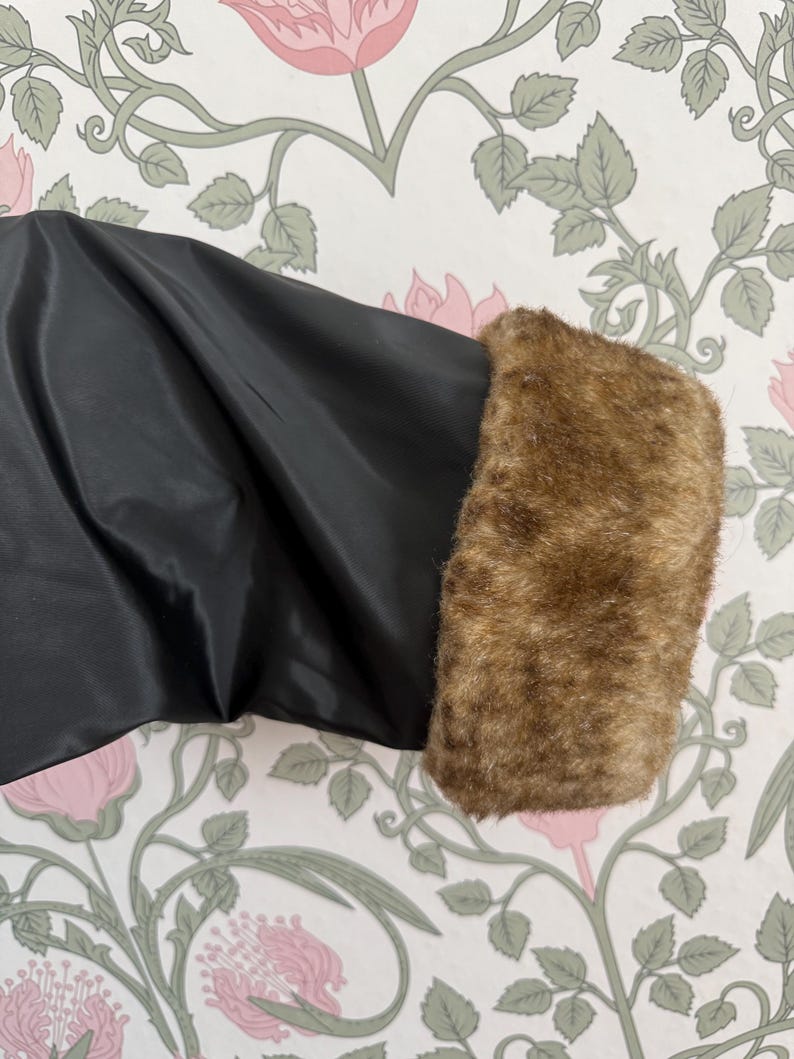 May include: Close-up of a black garment sleeve with a brown faux fur cuff. The sleeve is made of a shiny, smooth fabric. The faux fur cuff is a warm, light brown color, adding a touch of luxury. The background features a floral wallpaper.