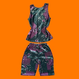 May include: A two-piece pajama set consisting of a sleeveless tank top and matching shorts. The fabric features a dark green and purple patchwork pattern with a plaid design, houndstooth accents, and scattered floral motifs throughout the garment.