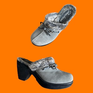 Pode incluir: Dois tamancos Skechers cinzentos com acabamento em pele sintética e um detalhe de laço. Um tamanco é mostrado de cima e o outro de lado. Os tamancos têm um salto e sola pretos. O logótipo Skechers é visível na palmilha.