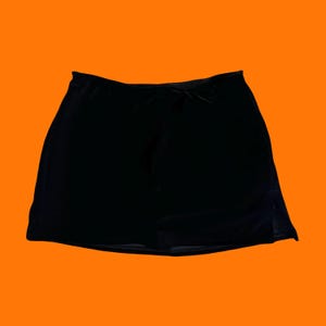 May include: A solid black mini skirt featuring a smooth, slightly reflective fabric finish. The garment has a simple, A-line silhouette with a clean, straight hemline and a subtle side slit detail, presented against a bright, solid orange background.