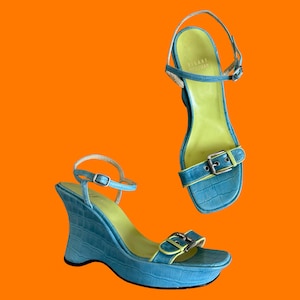 May include: Two blue wedge sandals with a textured pattern and lime green insoles. The sandals have ankle straps and buckle closures. The brand name "STUART WEITZMAN" is visible on the insole of one sandal.