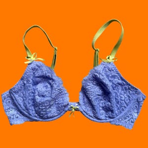 May include: A lavender lace bra with lime green straps and bows. The bra features a full coverage design with intricate floral lace detailing. The straps and bows add a pop of color against the lavender fabric. The bra is set against an orange background.