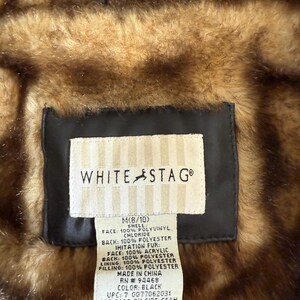 May include: Close-up of a White Stag jacket label. The label is rectangular with the brand name and a deer logo. The label provides details about the jacket's materials, including 100% polyvinyl chloride, polyester, and acrylic. The jacket is black and brown.