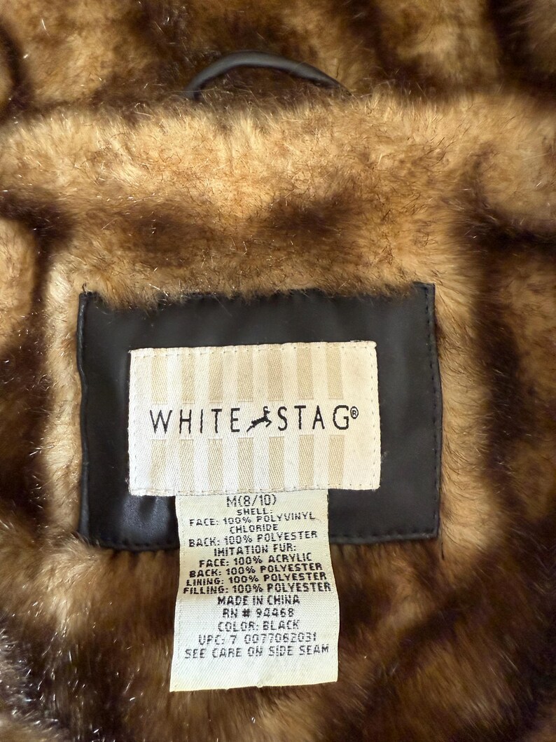 May include: Close-up of a White Stag jacket label. The label is rectangular with the brand name and a deer logo. The label provides details about the jacket's materials, including 100% polyvinyl chloride, polyester, and acrylic. The jacket is black and brown.