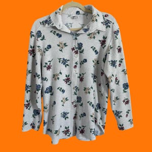 May include: A light gray button-down shirt with a floral pattern in blue, burgundy, and cream colors. The long-sleeved shirt has a collared neckline and is made of a ribbed fabric. The shirt is buttoned up and hanging on a beige hanger.
