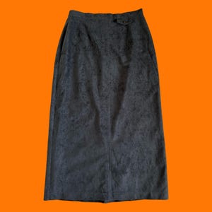 May include: A dark gray, knee-length skirt with a subtle, mottled pattern. The skirt features a button closure at the waistband and a back slit for ease of movement. The fabric appears to be a soft, suede-like material.