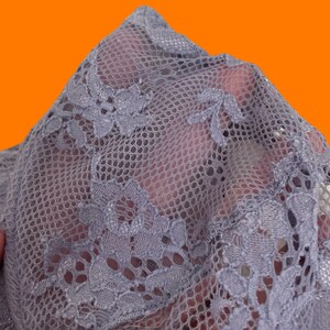 May include: Close-up of a sheer, gray lace fabric with a floral pattern. The delicate material is held up, revealing the intricate design and texture. The lace is a soft gray color, with a detailed floral motif.