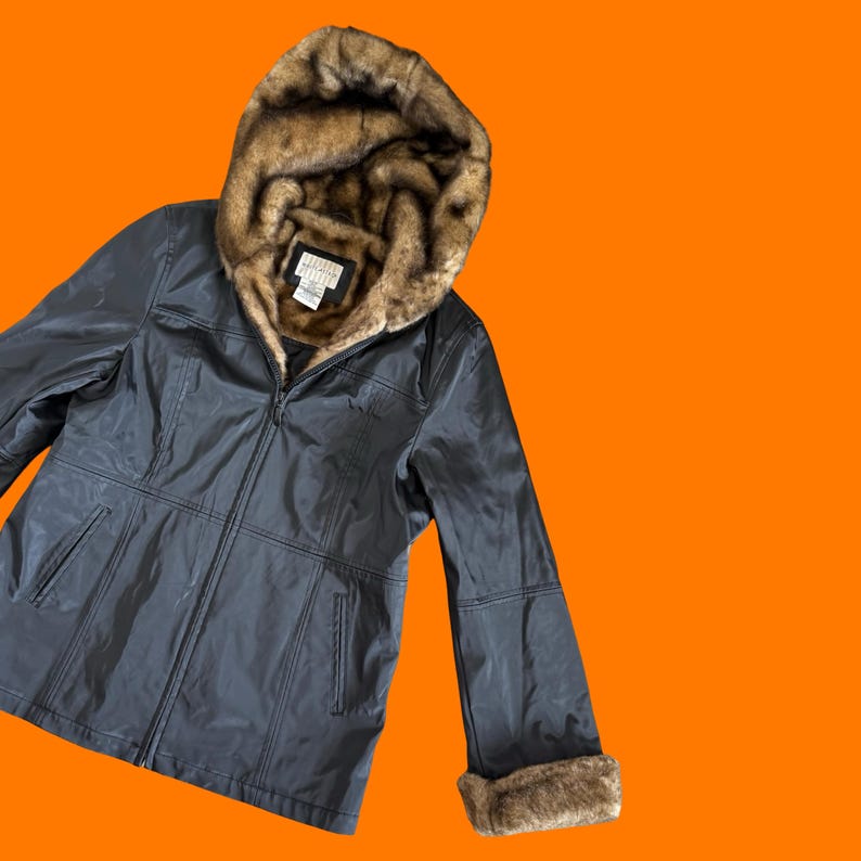May include: A dark gray jacket with a faux fur-lined hood and cuffs. The jacket has a zipper closure and a label on the inside of the hood. The jacket is set against an orange background.