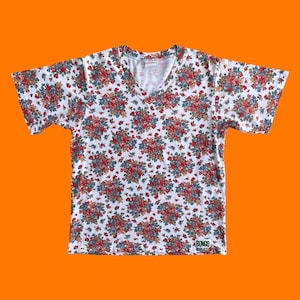 Op de afbeelding: Wit V-hals T-shirt met een bloemenpatroon. Het patroon heeft rode, blauwe en groene bloemen op een witte achtergrond. Het shirt heeft korte mouwen en een label met "BONGO".