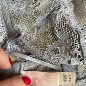 May include: A close-up of a light gray lace bra with a delicate floral pattern. The bra features a satin ribbon and a tag that reads "VICTORIA'S SECRET" and "34D". The bra is made of a soft, textured material.