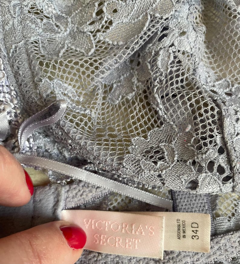 May include: A close-up of a light gray lace bra with a delicate floral pattern. The bra features a satin ribbon and a tag that reads "VICTORIA'S SECRET" and "34D". The bra is made of a soft, textured material.