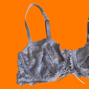 May include: A gray lace bra with adjustable straps and a delicate floral pattern. The bra features a corset-style front with white ribbon lacing and a small bow. The background is a solid orange.