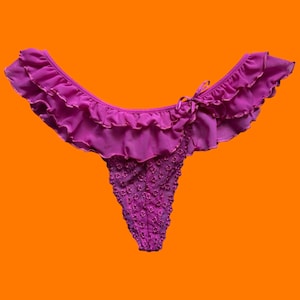 May include: A magenta thong with a ruffled trim and a small bow. The front of the thong is adorned with small, embroidered floral details. The background is a solid orange.