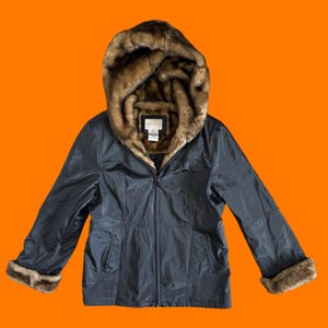 May include: A black zip-up jacket featuring a large hood lined with brown faux fur. The jacket includes long sleeves with matching brown faux fur cuffs and a central front zipper. A white rectangular brand label is visible inside the hood.
