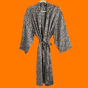 May include: A leopard print robe with a matching belt, hanging on a wooden hanger. The robe features a brown, black, and cream pattern. The robe is made of a soft, silky material and has wide sleeves.