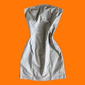 May include: A strapless, beige dress with a fitted bodice and a flared skirt. The dress has vertical seams and is made of a smooth fabric. The dress is set against a bright orange background.