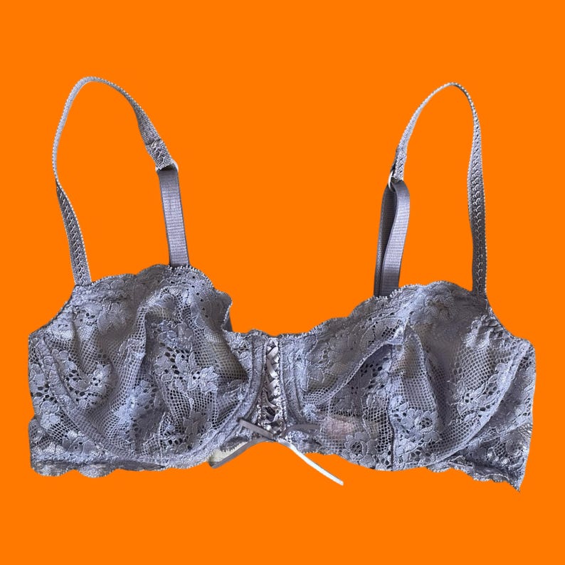 May include: A light gray lace bra with adjustable straps. The bra features intricate floral lace detailing and a small bow at the center. The background is a solid orange.