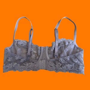 May include: A gray lace bra with adjustable straps and a hook-and-eye closure. The bra features a floral lace design along the edges and cups. The background is a solid orange.