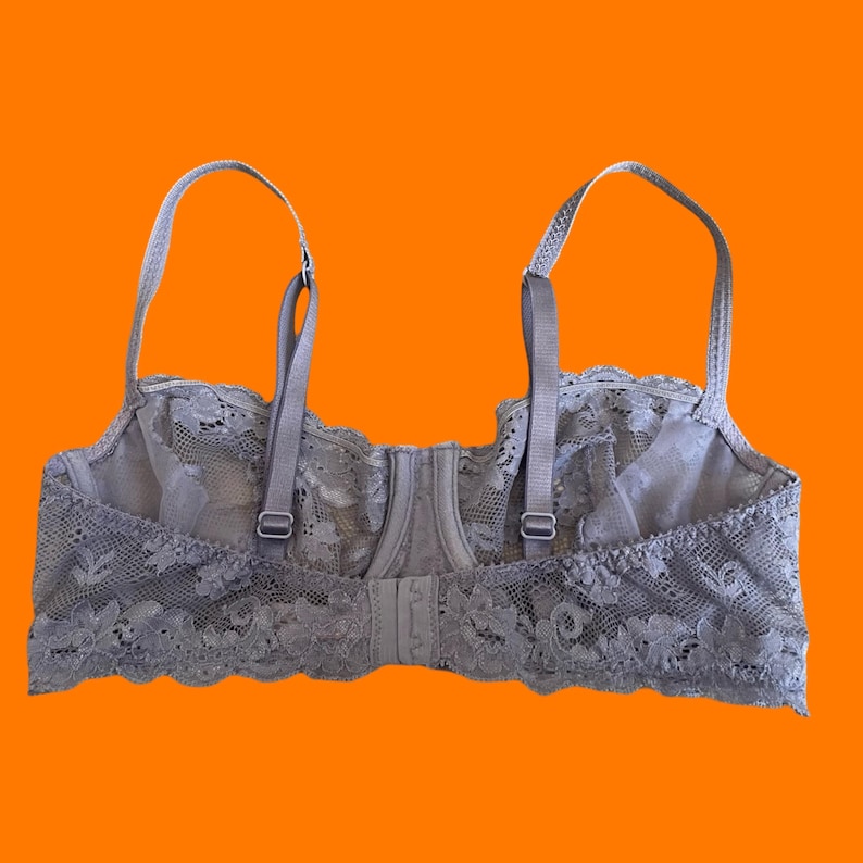May include: A gray lace bra with adjustable straps and a hook-and-eye closure. The bra features a floral lace design along the edges and cups. The background is a solid orange.