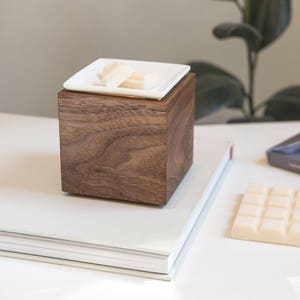 May include: A wooden cube diffuser with a white ceramic dish on top. The dish contains wax melts. The diffuser is sitting on a white book.