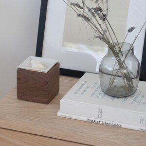 May include: A wooden cube with a white ceramic dish on top, a book titled "Charles & Ray Eames" by Taschen, and a glass vase with dried lavender stems on a light wood surface.