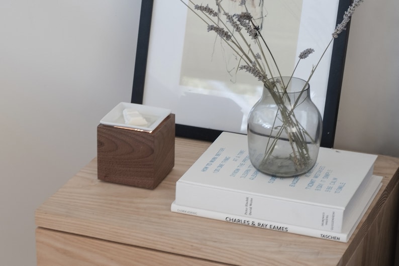 May include: A wooden cube with a white ceramic dish on top, a book titled "Charles & Ray Eames" by Taschen, and a glass vase with dried lavender stems on a light wood surface.