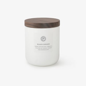 May include: White ceramic candle jar with a wooden lid. The jar has a label that reads "Black Ginger" and describes the scent as "worn leather and tobacco, touches of cedar, ginger and peppercorn."